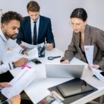 A diverse group of professionals in a business meeting, collaborating over documents and digital devices.
